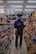 A person stands in the aisle of a store, browsing items on the shelves while looking at their phone. The shelves are filled with various snacks and packaged goods. Overhead fluorescent lights illuminate the space, and there is signage advertising discounts.
