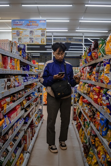 A person stands in the aisle of a store, browsing items on the shelves while looking at their phone. The shelves are filled with various snacks and packaged goods. Overhead fluorescent lights illuminate the space, and there is signage advertising discounts.