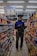 A person stands in the aisle of a store, browsing items on the shelves while looking at their phone. The shelves are filled with various snacks and packaged goods. Overhead fluorescent lights illuminate the space, and there is signage advertising discounts.
