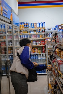 A person wearing a jacket and carrying a bag is browsing products in a store aisle. Shelves are stocked with a variety of goods, including drinks, snacks, and other grocery items. The setting appears to be a convenience or grocery store with bright lighting.