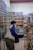 A person wearing a jacket and carrying a bag is browsing products in a store aisle. Shelves are stocked with a variety of goods, including drinks, snacks, and other grocery items. The setting appears to be a convenience or grocery store with bright lighting.