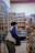 A person wearing a jacket and carrying a bag is browsing products in a store aisle. Shelves are stocked with a variety of goods, including drinks, snacks, and other grocery items. The setting appears to be a convenience or grocery store with bright lighting.