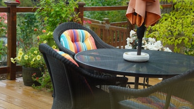 Round rattan patio dining table set with cushioned chairs and a portable umbrella base on a sunny deck.