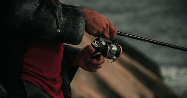 A person holding a fishing rod standing near a body of water. The focus is on the hands and the fishing reel, showcasing the grip and the details of the fishing equipment.