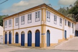A two-story colonial-style building with white walls and blue doors and window frames. The building has a yellow trim and a sloped roof. It's situated at the corner of a cobblestone street with some greenery in the background.