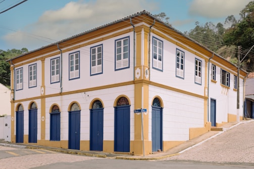 A two-story colonial-style building with white walls and blue doors and window frames. The building has a yellow trim and a sloped roof. It's situated at the corner of a cobblestone street with some greenery in the background.
