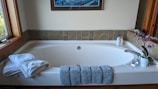 A clean and elegant bathroom features a large white bathtub with a modern design. At the front of the tub, there are neatly folded white towels and a textured blue bath mat draped over the edge. To the right, a potted orchid with purple flowers adds a touch of nature and color. Next to the orchid, two bottles of liquid soap or lotion are arranged near the faucet. The walls surrounding the tub are partially tiled with earthy-colored tiles, and a framed artwork hangs above the tub. A window on the left side lets natural light illuminate the space.