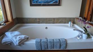 A clean and elegant bathroom features a large white bathtub with a modern design. At the front of the tub, there are neatly folded white towels and a textured blue bath mat draped over the edge. To the right, a potted orchid with purple flowers adds a touch of nature and color. Next to the orchid, two bottles of liquid soap or lotion are arranged near the faucet. The walls surrounding the tub are partially tiled with earthy-colored tiles, and a framed artwork hangs above the tub. A window on the left side lets natural light illuminate the space.