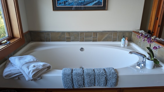 A clean and elegant bathroom features a large white bathtub with a modern design. At the front of the tub, there are neatly folded white towels and a textured blue bath mat draped over the edge. To the right, a potted orchid with purple flowers adds a touch of nature and color. Next to the orchid, two bottles of liquid soap or lotion are arranged near the faucet. The walls surrounding the tub are partially tiled with earthy-colored tiles, and a framed artwork hangs above the tub. A window on the left side lets natural light illuminate the space.