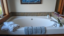 A clean and elegant bathroom features a large white bathtub with a modern design. At the front of the tub, there are neatly folded white towels and a textured blue bath mat draped over the edge. To the right, a potted orchid with purple flowers adds a touch of nature and color. Next to the orchid, two bottles of liquid soap or lotion are arranged near the faucet. The walls surrounding the tub are partially tiled with earthy-colored tiles, and a framed artwork hangs above the tub. A window on the left side lets natural light illuminate the space.