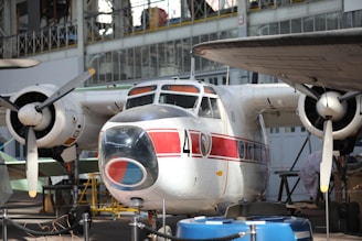 A vintage aircraft is on display inside an aviation museum. The aircraft has a distinct red and white paint scheme with the number 4 clearly visible on its fuselage. The propellers and engines are prominently featured, and the surrounding area includes museum ropes and other aviation equipment.
