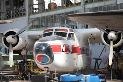 A vintage aircraft is on display inside an aviation museum. The aircraft has a distinct red and white paint scheme with the number 4 clearly visible on its fuselage. The propellers and engines are prominently featured, and the surrounding area includes museum ropes and other aviation equipment.