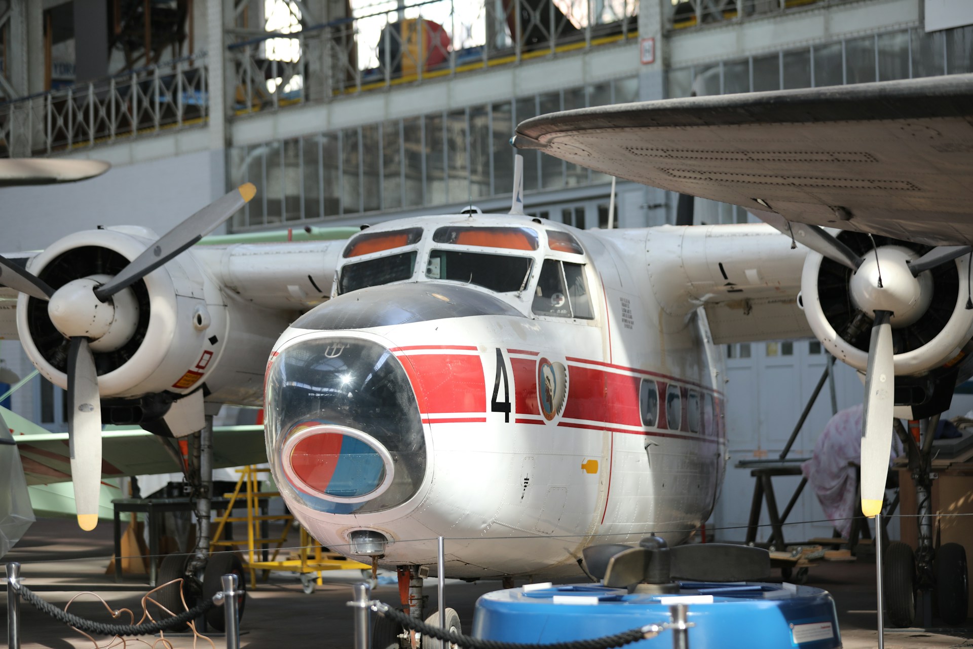 A vintage aircraft is on display inside an aviation museum. The aircraft has a distinct red and white paint scheme with the number 4 clearly visible on its fuselage. The propellers and engines are prominently featured, and the surrounding area includes museum ropes and other aviation equipment.