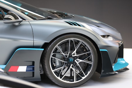 A close-up shot of a luxury sports car wheel and part of the body, showcasing intricate design details. The car features a sleek metallic finish with blue and black accents. The wheel is large, with a multi-spoke design and the Bugatti emblem in the center, indicating high performance engineering.
