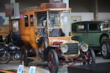 A vintage car with a wooden body and brass detailing is prominently displayed. The intricate design of the car includes a leather seat and an open top. In the background, other classic cars, including a green antique car and a motorcycle, are visible in a showroom setting. The lighting highlights the polished surfaces of the vehicles, and there's a historical ambiance with posters and framed images on the walls.