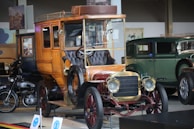 A vintage car with a wooden body and brass detailing is prominently displayed. The intricate design of the car includes a leather seat and an open top. In the background, other classic cars, including a green antique car and a motorcycle, are visible in a showroom setting. The lighting highlights the polished surfaces of the vehicles, and there's a historical ambiance with posters and framed images on the walls.