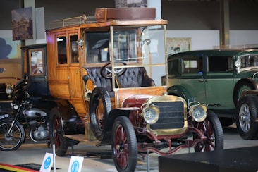 A vintage car with a wooden body and brass detailing is prominently displayed. The intricate design of the car includes a leather seat and an open top. In the background, other classic cars, including a green antique car and a motorcycle, are visible in a showroom setting. The lighting highlights the polished surfaces of the vehicles, and there's a historical ambiance with posters and framed images on the walls.