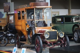 A vintage car with a wooden body and brass detailing is prominently displayed. The intricate design of the car includes a leather seat and an open top. In the background, other classic cars, including a green antique car and a motorcycle, are visible in a showroom setting. The lighting highlights the polished surfaces of the vehicles, and there's a historical ambiance with posters and framed images on the walls.