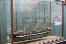 A detailed model of a classic sailing ship encased in a glass display. The ship features multiple masts with intricate rigging and a hull painted with dark blue and green. The model is displayed on a wooden base within a glass case.