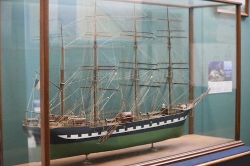 A detailed model of a classic sailing ship encased in a glass display. The ship features multiple masts with intricate rigging and a hull painted with dark blue and green. The model is displayed on a wooden base within a glass case.