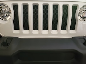 A close-up view of the front grille of a vehicle, featuring vertical slats and two round headlights. The grille is white with black mesh, and the bumper is dark gray.