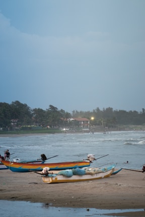 Several fishing boats are resting on a sandy beach with waves crashing in the background. The sky is overcast, and the coastline is lined with palm trees and buildings in the distance. A few people can be seen near the water and on the beach.