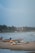 Several fishing boats are resting on a sandy beach with waves crashing in the background. The sky is overcast, and the coastline is lined with palm trees and buildings in the distance. A few people can be seen near the water and on the beach.
