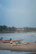 Several fishing boats are resting on a sandy beach with waves crashing in the background. The sky is overcast, and the coastline is lined with palm trees and buildings in the distance. A few people can be seen near the water and on the beach.