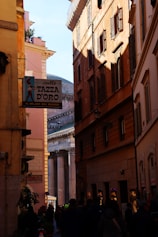 A narrow street flanked by tall historical buildings, some lit by sunlight and others in shadow. A sign for 'Caffè Tazza d'Oro' is visible on one side, and at the end of the street, there is an ancient structure with columns. People are walking along the street, and storefronts can be seen on the right side.
