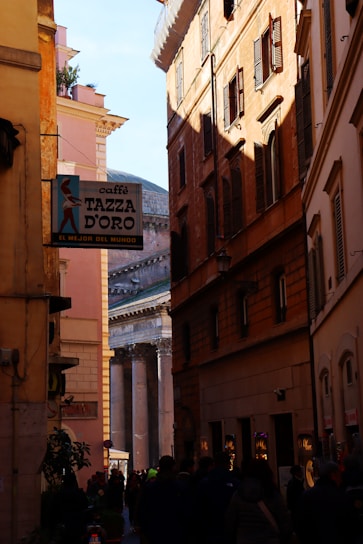A narrow street flanked by tall historical buildings, some lit by sunlight and others in shadow. A sign for 'Caffè Tazza d'Oro' is visible on one side, and at the end of the street, there is an ancient structure with columns. People are walking along the street, and storefronts can be seen on the right side.