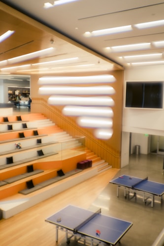 A modern interior space featuring a series of tiered, orange-cushioned seating with black pillows and a wooden staircase. In front of the seating area are two blue ping pong tables. The room has high ceilings with a unique lighting design and a large screen mounted on one wall.
