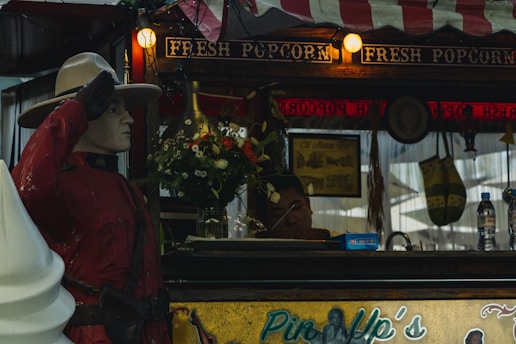 A popcorn stall adorned with vibrant lights and a sign reading 'Fresh Popcorn' features a Mountie statue in the foreground, saluting. A bouquet of flowers is arranged on the counter, where a staff member appears to be resting. Surrounding elements include retro decor and bottled water on a shelf.