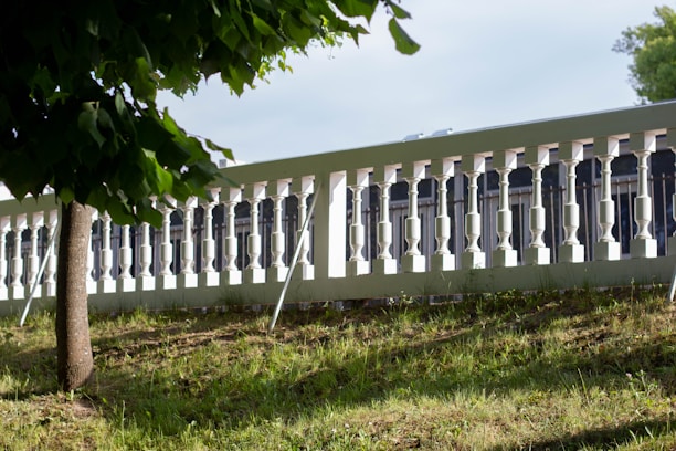 A well-maintained grassy area with a small tree casting a shadow in the foreground. Behind the tree is a white decorative fence with evenly spaced vertical balusters, extending across the image.