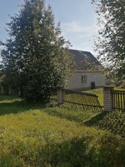 A house with a sloped roof is partially hidden behind tall, leafy trees. The house is made of light-colored bricks. A wooden fence runs along the property, some parts of which are slightly tilted, and the grass in the yard is lush and green.