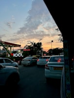A busy road scene during sunset, with multiple cars lined up in traffic in front of an Indian Oil fuel station. The sky above is partly cloudy, displaying a gradient of colors from orange to blue. Trees and streetlights are scattered along the roadside.