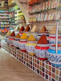 A vibrant spice shop interior featuring an array of large, ornate ceramic jars filled with colorful spices, each forming small mounds. The jars are decorated with intricate blue and red patterns. Shelves in the background hold various packaged goods and spices, contributing to the colorful and lively atmosphere of the shop.