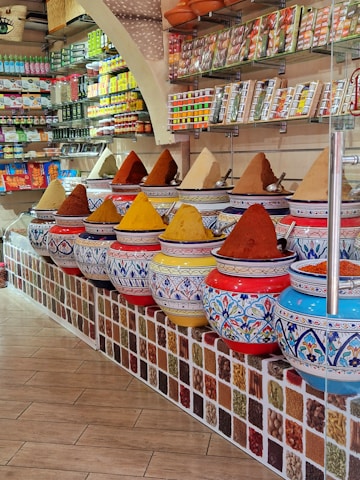 A vibrant spice shop interior featuring an array of large, ornate ceramic jars filled with colorful spices, each forming small mounds. The jars are decorated with intricate blue and red patterns. Shelves in the background hold various packaged goods and spices, contributing to the colorful and lively atmosphere of the shop.