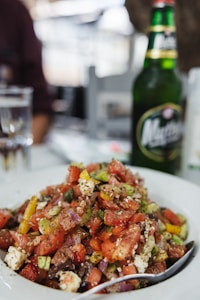 A plate of salad featuring a mix of chopped vegetables like tomatoes, cucumbers, peppers, red onion, and feta cheese, garnished with herbs. A green glass bottle of beer is positioned next to the salad, along with a glass partially filled with water. The setting appears to be outdoors or in a casual dining environment.