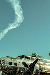A vintage military airplane with a polished metal exterior is pictured against a clear blue sky. The plane features nose art depicting a pin-up girl, and the airframe has multiple panels and rivets visible. A smoke trail curls through the sky overhead.