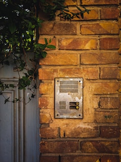 A weathered metal intercom installed on a rustic brick wall, with lush green leaves partially covering the top left area. The intercom features several vents and buttons, framed by the aged texture of the bricks.