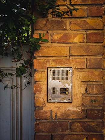A weathered metal intercom installed on a rustic brick wall, with lush green leaves partially covering the top left area. The intercom features several vents and buttons, framed by the aged texture of the bricks.