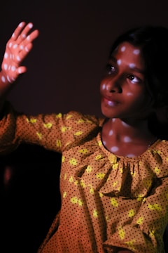 A girl with a pensive expression gazes upwards, her hand raised against the light. She wears a yellow polka dot dress, and the light casts a pattern of bright spots on her face and clothing, creating an interesting play of shadows and highlights.