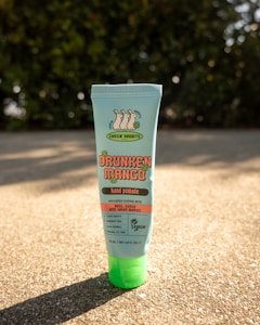A tube of hand pomade labeled 'Chasin' Rabbits Drunken Mango' is standing upright on a sunlit surface. The product is described as being delicately scented with basil, guava, and green mango. It highlights attributes such as being clean beauty, paraben-free, vegan, and mineral oil-free. The packaging is primarily light blue with orange and green accents. The background features a blurred view of greenery.