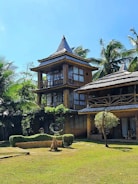 A rustic multi-story wooden house with a thatched roof is nestled among tall palm trees. The structure features large windows and a natural, earthy design. The surrounding area has well-maintained greenery, including a lawn and sculpted bushes, contributing to a tranquil and tropical atmosphere under a clear blue sky.