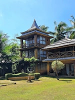 A rustic multi-story wooden house with a thatched roof is nestled among tall palm trees. The structure features large windows and a natural, earthy design. The surrounding area has well-maintained greenery, including a lawn and sculpted bushes, contributing to a tranquil and tropical atmosphere under a clear blue sky.