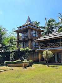 A rustic multi-story wooden house with a thatched roof is nestled among tall palm trees. The structure features large windows and a natural, earthy design. The surrounding area has well-maintained greenery, including a lawn and sculpted bushes, contributing to a tranquil and tropical atmosphere under a clear blue sky.