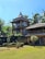 A rustic multi-story wooden house with a thatched roof is nestled among tall palm trees. The structure features large windows and a natural, earthy design. The surrounding area has well-maintained greenery, including a lawn and sculpted bushes, contributing to a tranquil and tropical atmosphere under a clear blue sky.