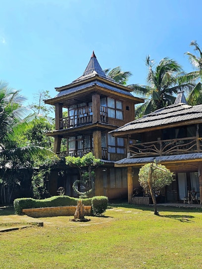 A rustic multi-story wooden house with a thatched roof is nestled among tall palm trees. The structure features large windows and a natural, earthy design. The surrounding area has well-maintained greenery, including a lawn and sculpted bushes, contributing to a tranquil and tropical atmosphere under a clear blue sky.