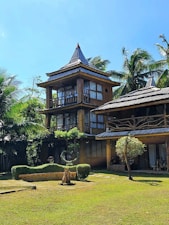 A rustic multi-story wooden house with a thatched roof is nestled among tall palm trees. The structure features large windows and a natural, earthy design. The surrounding area has well-maintained greenery, including a lawn and sculpted bushes, contributing to a tranquil and tropical atmosphere under a clear blue sky.