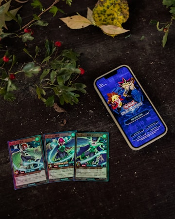 A smartphone displaying the Yu-Gi-Oh! Duel Links game title screen is placed on a dark surface surrounded by leaves and red berries. Below the phone, there are three Yu-Gi-Oh! trading cards laid out, depicting different characters.